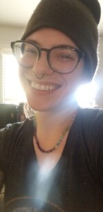 a person with light skin, wearing round black frame glasses,a grey vneck shirt and a rainbow crystal necklace with a septum ring, a monroe piercing and a grey cap smiles